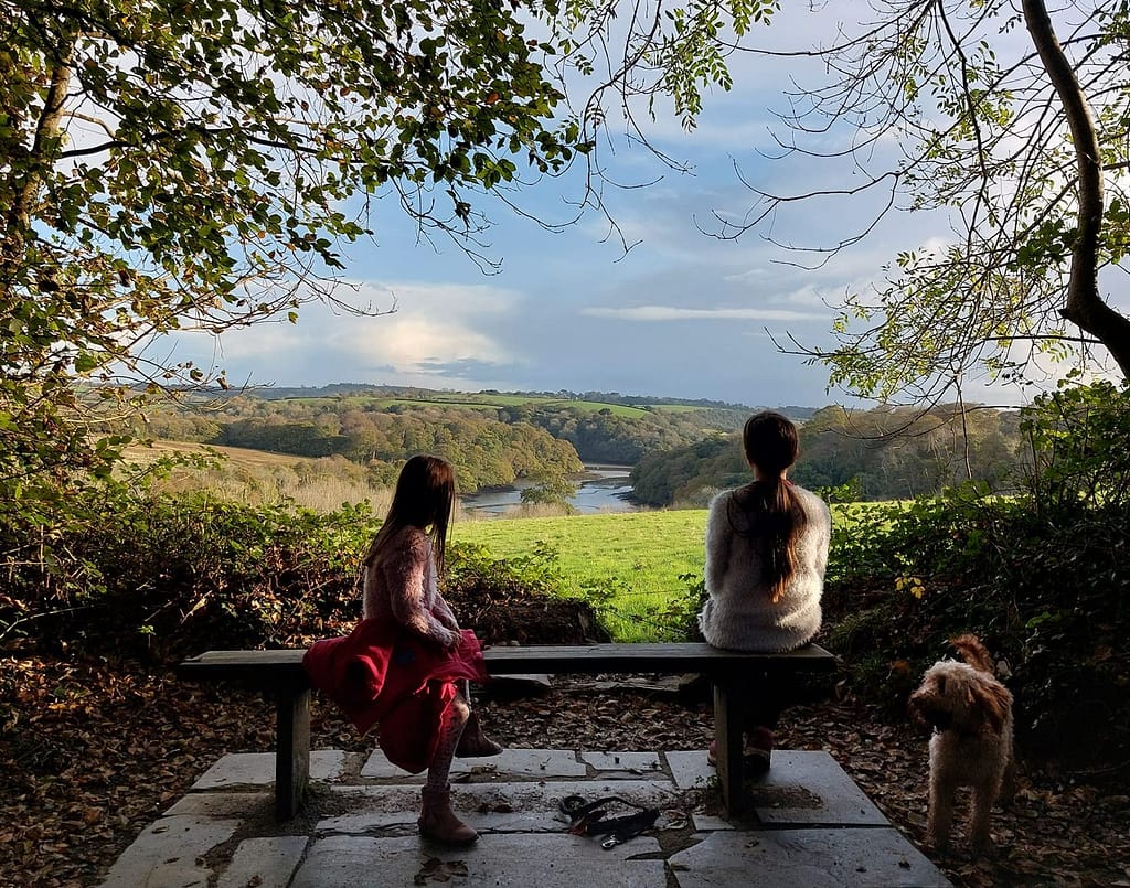 Two people and a dog sitting on a bench beneath autumn trees, looking out over a peaceful countryside view — a moment of calm and connection with nature for mental health day 2025