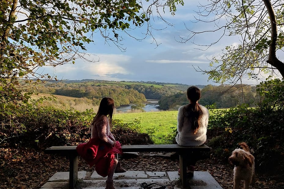 Two people and a dog sitting on a bench beneath autumn trees, looking out over a peaceful countryside view — a moment of calm and connection with nature for mental health day 2025